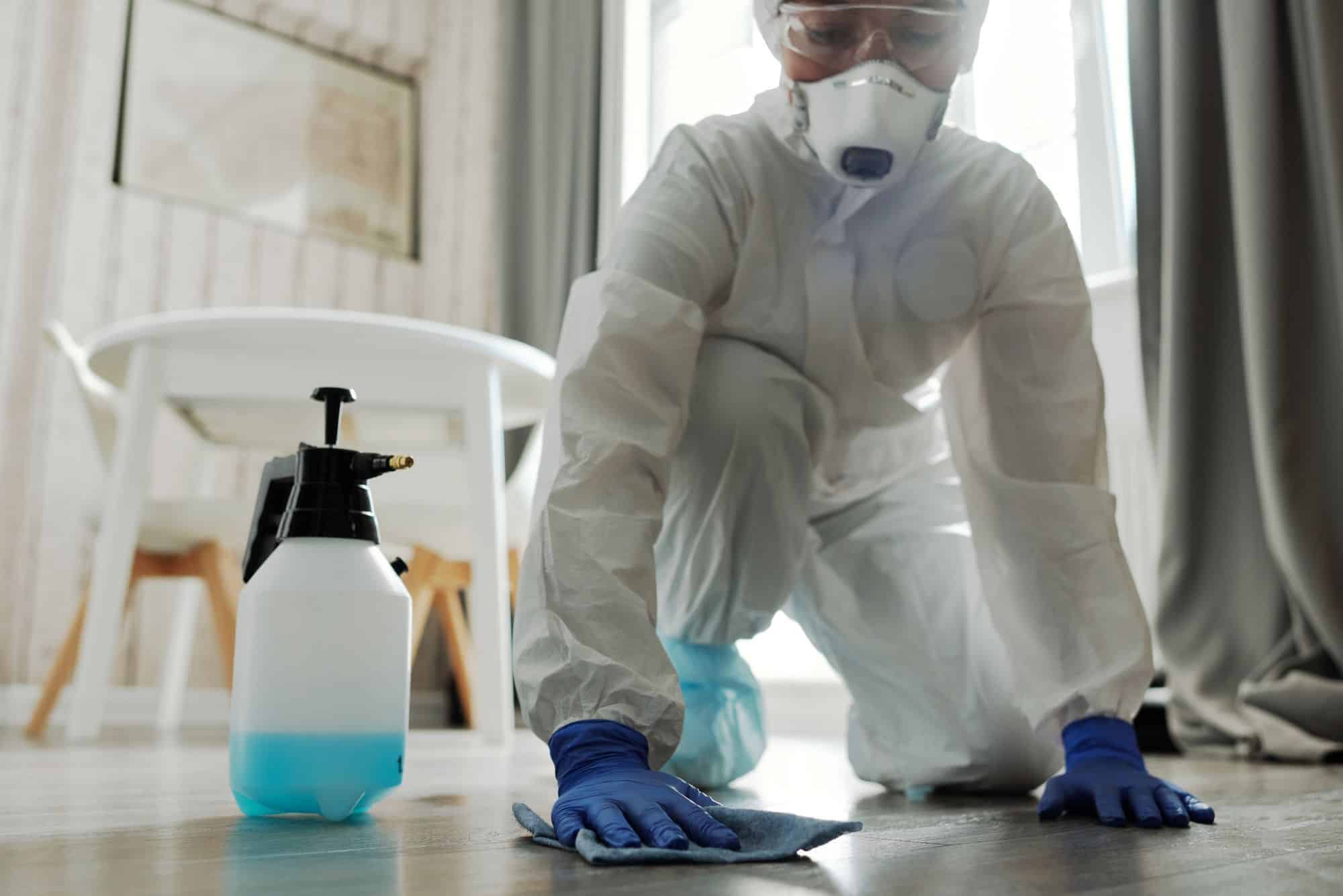 Mould remediation technician in a protective suit and mask wiping down a floor with antimicrobial treatment